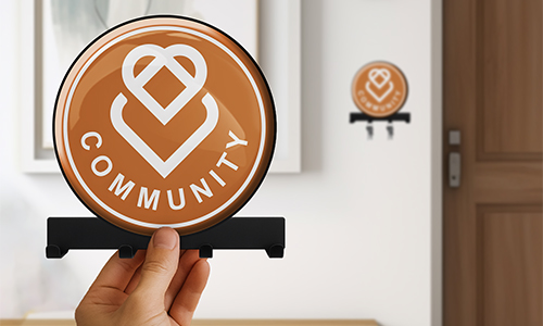 Hand hält rundes COMMUNITY Schild mit Herz-Logo, orange Hintergrund; gleiches Schild an Wand neben Tür