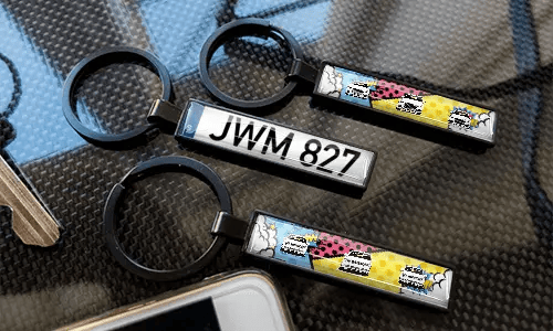 Sarjakuvatyyliset rekisterikilpi-avaimenperät, esillä hiilikuitutekstuurisella pinnalla värikkäillä autodesigneilla