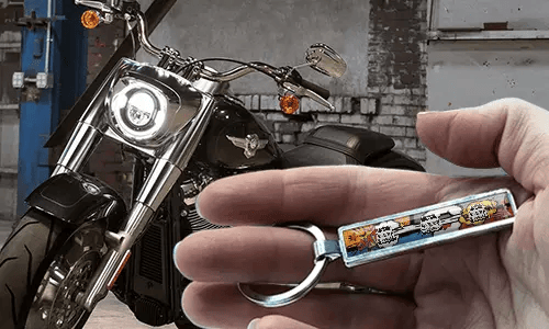 Klíčenka s poznávací značkou v komiksovém stylu, držená v ruce, s motocyklem v pozadí