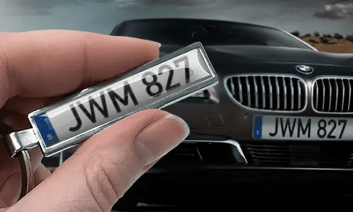 Portachiavi a forma di targa in stile fumetto con numero personalizzato, tenuto in mano vicino alla targa dell’auto corrispondente